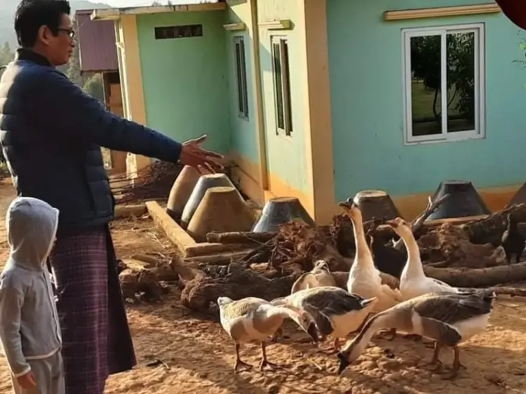 Ein Mann zeigt auf Gänse vor einem hellgrünen Haus.