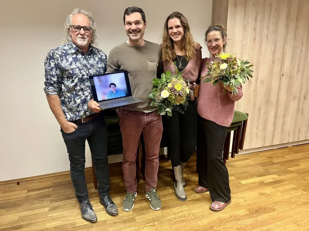 Vier Vorstandsmitglieder stehen lächelnd nebeneinander, zwei Frauen halten Blumensträuße. Der Mann links hält einen Laptop, auf dem Marc Heintz per Videokonferenz zu sehen ist.