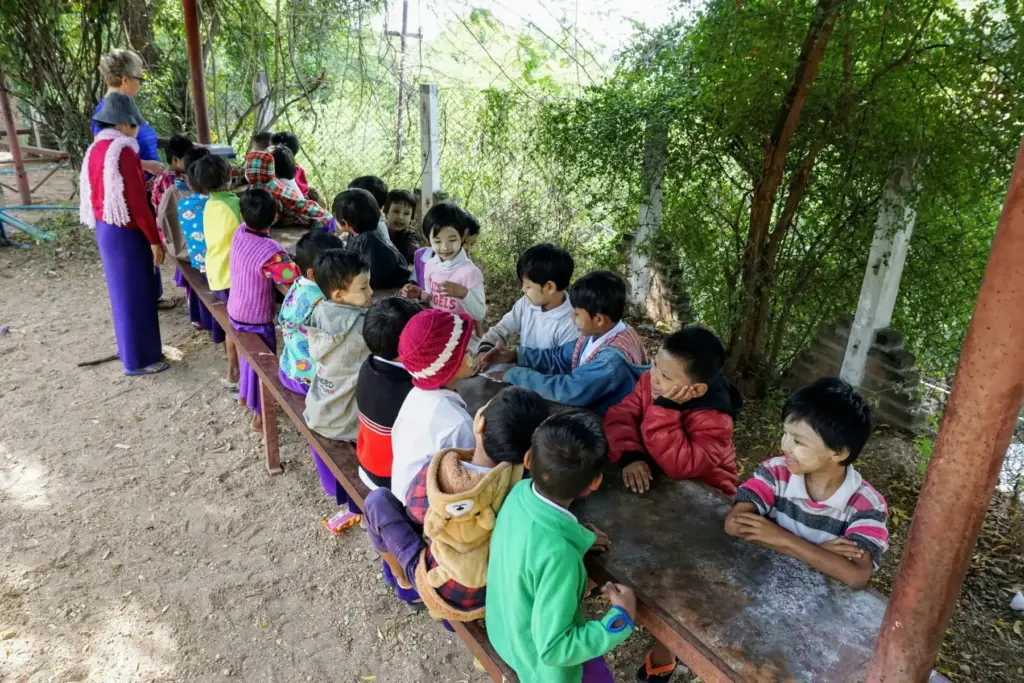 Kinder sitzen an einem langen Tisch im Freien unter Bäumen.