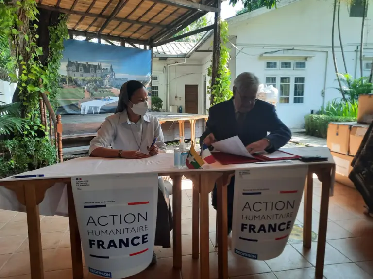 Zwei Personen unterzeichnen an einem Tisch mit Plakaten der Action humanitaire France.