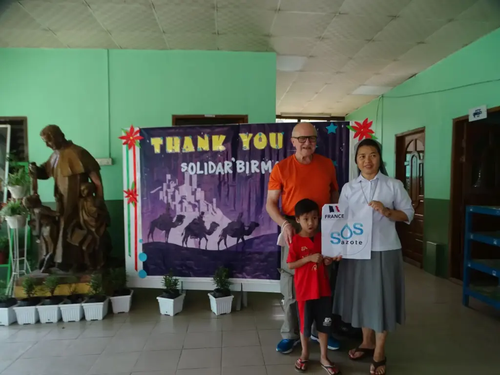 Ein Besucher, eine Frau und ein Kind vor einem Plakat mit der Aufschrift Thank you Solidar Birmanie.