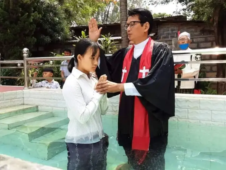 Ein Mädchen wird im Wasserbecken getauft, der Pastor hebt segnend die Hand.